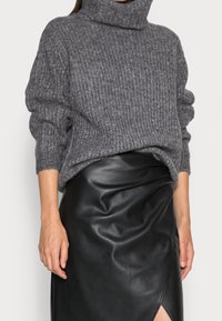 Kvinna som bär en grå grovstickad polotröja och en svart läderskjort med en slits framtill, stående mot en enkel bakgrund.