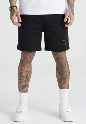 Persona con pantalones cortos negros con un pequeño emblema bordado, calcetines blancos, zapatillas deportivas blancas y tatuajes que cubren ambas piernas y manos.