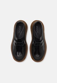 Une paire de chaussures en cuir noir à enfiler avec des semelles épaisses brunes, des orteils arrondis et des semelles intérieures avec logo, présentée en vue de dessus sur un fond clair.