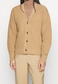 Beige neulecardigan, jossa on v-pääntie, napitettu etuosa, ribattuja hihansuita ja teksturoitu kuvio. Korostettu logolla hihassa.