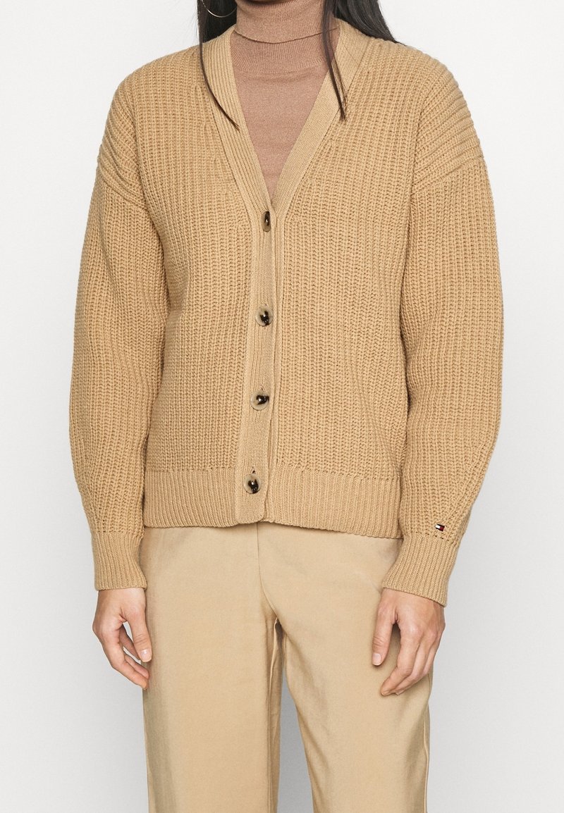 Beige neulecardigan, jossa on v-pääntie, napitettu etuosa, ribattuja hihansuita ja teksturoitu kuvio. Korostettu logolla hihassa.