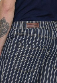 Pantalons en denim rayés avec une base bleu foncé et des lignes blanches verticales. Présente une étiquette en cuir et des coutures visibles sur les poches.