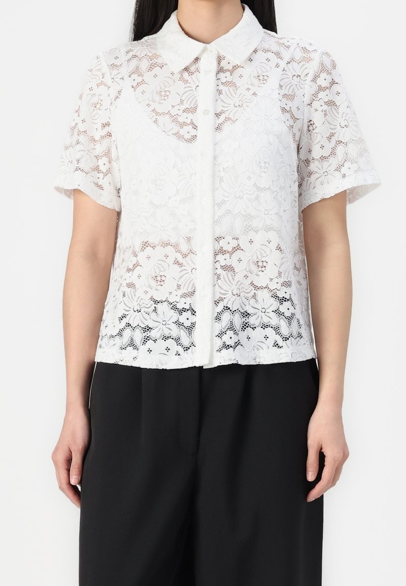 Femme portant une chemise blanche en dentelle à manches courtes avec des motifs floraux et un pantalon noir à jambes larges sur fond blanc.