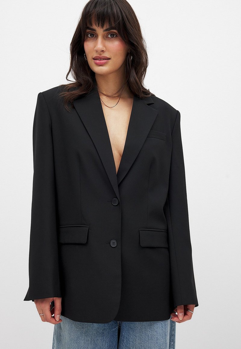 Schwarzer Blazer aus glattem Stoff, mit tiefem V-Ausschnitt, zwei Fronttaschen und zwei Knöpfen, sowie leicht übergroßen Ärmeln.