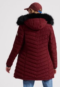 Maronfarvet pufferjakke med hætte, der har en faux fur kant, chevron quilting og sidelommer. Bagsiden har en buet kant og tekstureret materiale.
