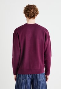 Felpa viola con colletto tondo, polsini e orlo a costine. Presenta una vestibilità rilassata. Indossata con pantaloni larghi a righe blu.