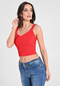Rotes, kurzes Oberteil mit V-Ausschnitt und breiten Trägern. Aus dehnbarem Material gefertigt und verfügt über eine kleine Tasche vorne. Kombiniert mit blauen Jeans.