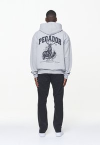 Grå hoodie med en stor svart grafik som föreställer en hjort och text, kombinerad med svarta byxor och vita sneakers. Slät tyg, avslappnad passform.