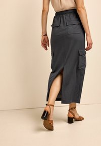 Jupe midi grise avec taille élastique, poches latérales et une fente à l'arrière. Associée à des sandales à talons couleur cuir clair. Texture lisse, design décontracté.