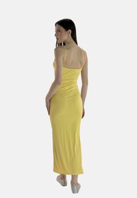 Abito maxi giallo, aderente e senza maniche, con scollatura a barca, realizzato in tessuto morbido. Il modello indossa scarpe piatte decorate, che mostrano una texture liscia.