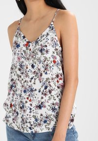 Top in tessuto leggero con stampa floreale. Presenta una scollatura a V, spalline sottili e uno sfondo bianco con fiori colorati in rosso, blu e verde.
