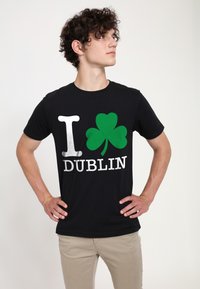 Mladý muž s kudrnatými vlasy, oblečený v černém tričku s nápisem "I" a zeleným trojlístkem a "DUBLIN", stojící s rukama na bocích a dívající se na stranu.