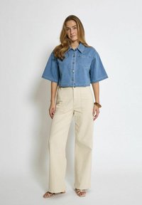 Kurze, blaue Jeansjacke mit kurzen Ärmeln, einer Frontknopfschließe und Tasche, kombiniert mit weißen, weiten Hosen.