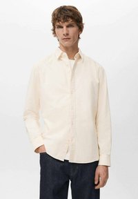 Camicia a maniche lunghe color crema, in cotone, con colletto classico e taglio dritto, indossata sopra una T-shirt bianca.