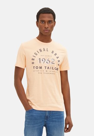 Pfirsichfarbener Baumwoll-T-Shirt mit Rundhalsausschnitt, kurzen Ärmeln und grafischem Frontdruck, einschließlich "Original Brand" und "1962" in dunkler Schrift.