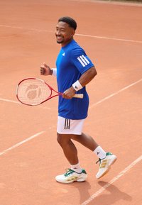 Kék sportpóló fehér csíkokkal, fehér rövidnadrág, fehér cipő zöld részletekkel. Egy piros teniszütőt tart a teniszpályán.