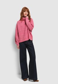 Roze, oversized, gebreid trui met een hoge hals, gecombineerd met donkerblauwe, wijde jeans. De outfit heeft een ontspannen pasvorm.