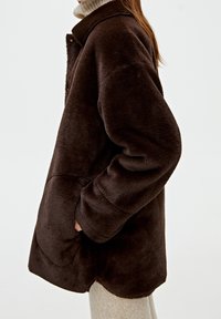 Brown fleece jacket with a collared neckline, side pockets, and a curved hem, featuring a soft, textured surface.