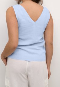 Top sans manches bleu clair avec un dos en V, fabriqué à partir d'un tissu texturé, associé à un pantalon de couleur claire et ample.