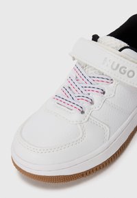 Baskets blanches en cuir texturé, avec une semelle en gomme, des lacets rayés rouges et noirs, et une sangle velcro avec la marque Hugo.