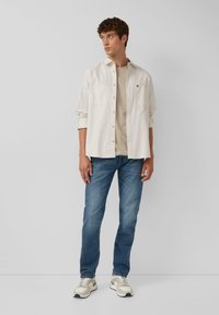 Lichtbeige button-up shirt over een neutraal T-shirt, gecombineerd met blauwe jeans en beige sneakers. Katoenen materiaal en een casual pasvorm.