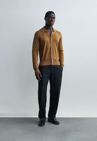 Άνδρας που στέκεται μπροστά σε έναν απλό γκρι τοίχο, φορώντας καφέ ζακέτα με κουμπιά, μαύρο παντελόνι κομμένο στα μέτρα και μαύρα παπούτσια φόρμας.