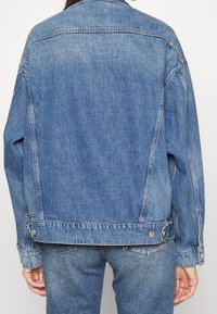 Chaqueta de denim azul claro con un corte relajado, costuras visibles y acentos de botones plateados. Presenta un collar clásico y grandes bolsillos traseros.