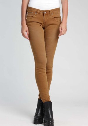 Osoba nosząca brązowe dopasowane spodnie, biały top i czarne botki, stojąca na tle jednolitego jasnego tła.