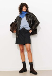 Femme debout, portant une veste en cuir noire, un pull bleu, une écharpe à motifs bleus, une jupe noire boutonnée, des bottes noires, et tenant un sac marron.