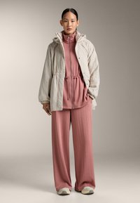 Femme portant un haut zippé rose poudré et un pantalon large avec une veste matelassée beige et des baskets claires, debout devant un fond uni.