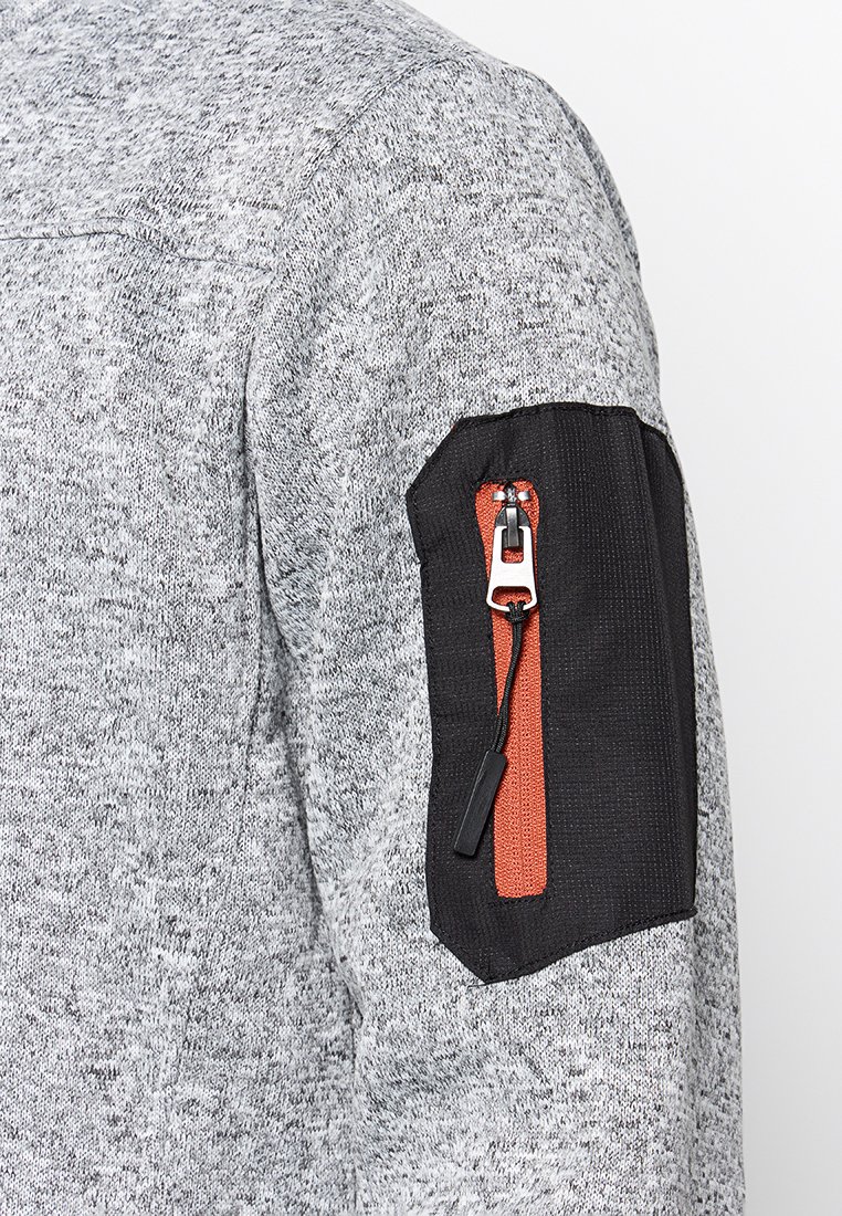 Graue strukturierte Strickjacke mit einem schwarzen Stoffärmeln, der eine orangefarbene Reißverschlusstasche hat. Die Tasche ist mit einem silbernen Reißverschluss-Zipper ausgestattet.