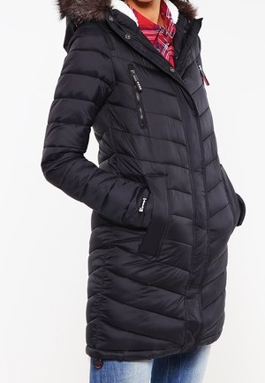 Kobieta w długiej czarnej kurtce puchowej z kapturem obszytym futerkiem i w czerwonej koszuli w kratę, z rękami w bocznych kieszeniach, na białym tle.