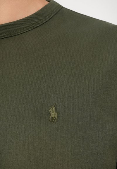 Olive krāsas t-krekls, kas izgatavots no mīksta kokvilnas, ar ribotu apaļu kakla izgriezumu un maigu izšuvumu kreisajā krūtī.
