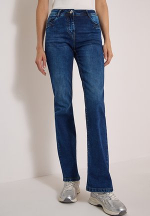Femme portant un jean droit bleu foncé et des baskets blanches, debout devant un fond clair.