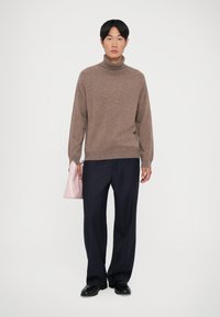 Brown wool turtleneck sweater, relaxed fit, paired with navy pinstriped trousers. Model holds a light pink handbag. Black shoes complete the look.