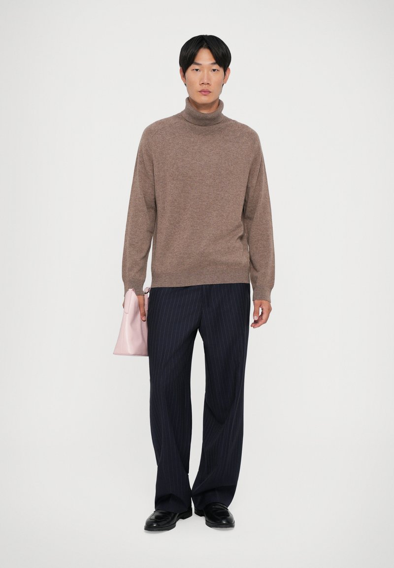 Pull en laine marron à col roulé, coupe décontractée, associé à un pantalon bleu marine à rayures. Le mannequin tient un sac à main rose clair. Des chaussures noires complètent le look.