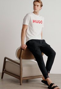 Bílý tričko s červeným potiskem "HUGO", spojené s černými tepláky a černými nazouvacími sandály, sedí na světlé dřevěné židli.