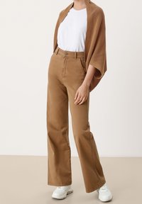 Bruine broek met wijde pijpen en hoge taille, voorzien van knoopsluiting en steekzakken aan de voorkant; gecombineerd met een los wit T-shirt en een bruine gebreide sjaal.