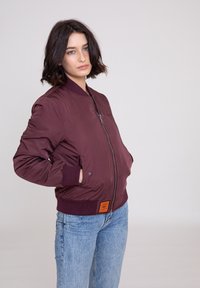 Bordeaux bomberjakke med lynlås foran, ribbede manchetter og et orange mærke. Har en glat overflade og en cropped design.