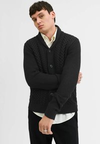 Dunkel strukturierte Strickjacke mit breitem Schalkragen, Zopfdesign und Knopfverschluss. Getragen über einem hellen Hemd mit sichtbarem Bündchen.