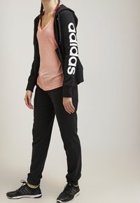 Veste à capuche zippée noire avec le logo "adidas" blanc sur la manche, t-shirt rose à col en V, pantalon de survêtement noir et chaussures de sport noires avec semelles blanches.