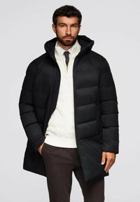 Giacca a vento nera con cappuccio, dotata di chiusura con zip. Indossata su un maglione bianco a maglia cable e pantaloni marroni. Texture liscia, motivo trapuntato.
