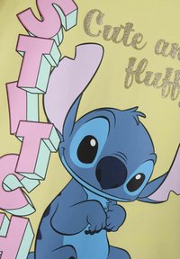 Żółta koszulka z krótkim rękawem z niebieską postacią z kreskówki z dużymi uszami, tekstem w kolorze różowym i błyszczącymi złotymi akcentami.