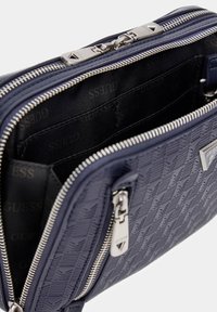 Geantă din piele matlasată de culoare bleumarin, cu fermoar și accesorii din argint. Interiorul are un căptușeală din material închis la culoare, cu un model repetat al logo-ului "Guess".