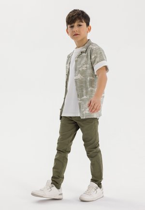 Junge trägt ein kurzärmliges, hellgrünes gemustertes Hemd über einem weißen T-Shirt, kombiniert mit olivgrünen Hosen und weißen Sneakers. Einfaches Design.