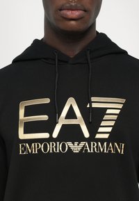 Fekete kapucnis pulóver arany nyomtatott "EA7 EMPORIO ARMANI" felirattal és logóval. Zsinóros kapucnival és bordázott textúrával rendelkezik.