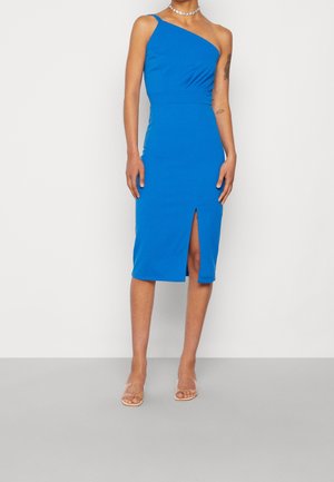 Femme portant une robe midi bleu vif à une épaule avec une fente à l'avant et des sandales à talons hauts transparentes sur un fond uni.