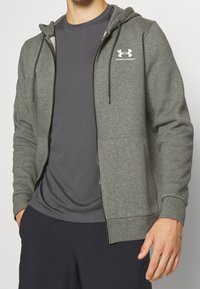 Felpa grigia con zip e cappuccio regolabile, tasche laterali e logo bianco sul petto. Indossata sopra una maglietta a girocollo grigio scuro.