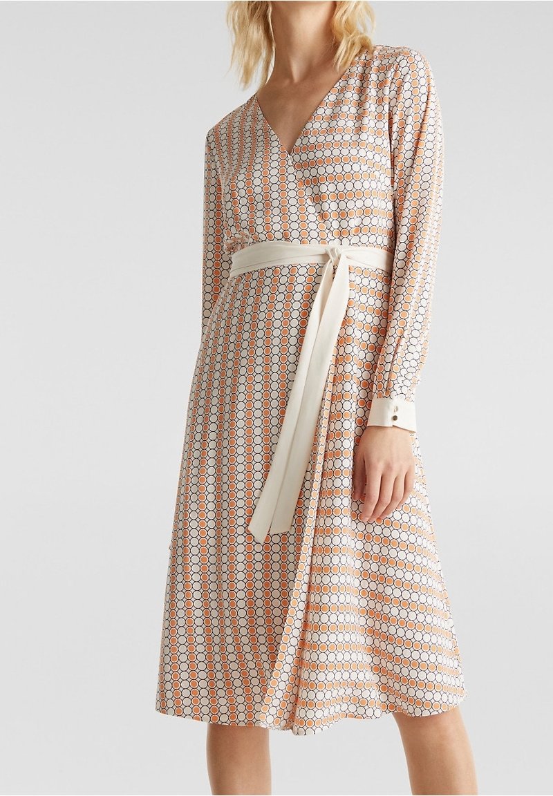 Femme portant une robe portefeuille mi-longue à manches longues avec un motif circulaire orange, blanc et noir, ceinturée d'une large ceinture crème à la taille.