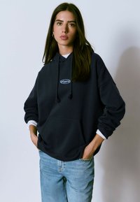 Sudadera con capucha azul marino, con un bolsillo frontal, cordones ajustables y un pequeño logo. Combinada con jeans azul claro.